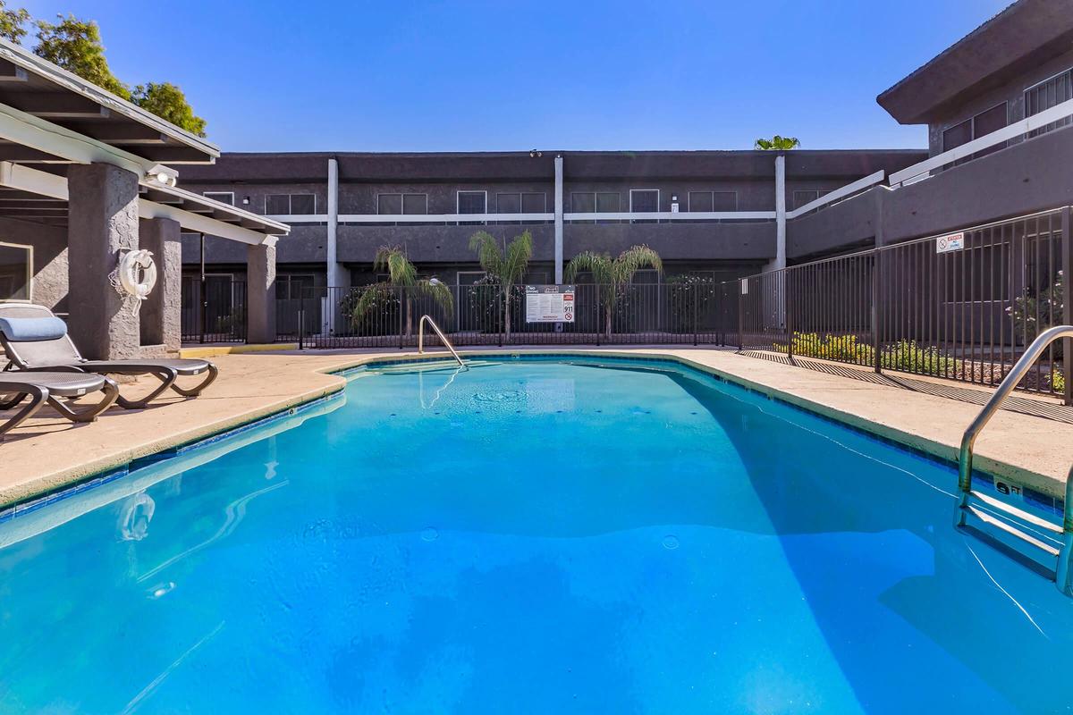 Bright blue swimming pool surrounded by lounge chairs and lush greenery, with modern apartments in the background. The sky is clear and sunny, creating a relaxing outdoor atmosphere. A safety sign is posted near the pool.