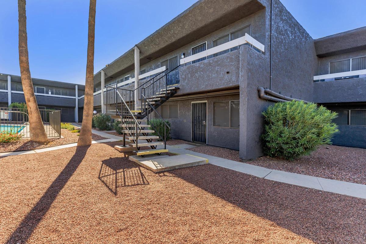 A two-story apartment building with a staircase leading to the second floor. The exterior features dark gray walls and white railings. Surrounding the building are palm trees and landscaped gravel areas. A swimming pool is visible in the background, along with a fenced area.