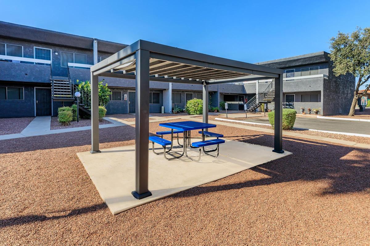 A modern outdoor seating area featuring a blue picnic table under a black metal pergola, surrounded by gravel and landscaped with bushes. In the background, apartment buildings with staircases are visible against a clear blue sky.