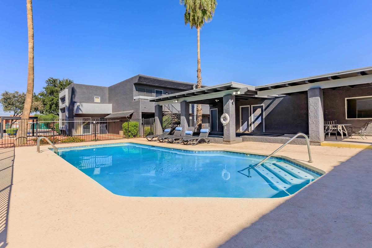 A clear blue swimming pool surrounded by lounge chairs, with a palm tree in the background. The pool area is enclosed with a fence, and there are buildings nearby. The sky is bright and clear, indicating a sunny day.