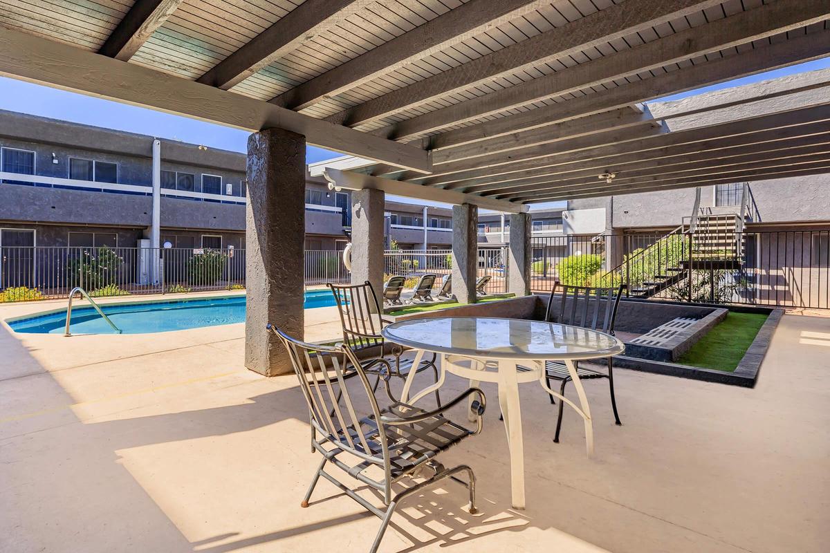A sunny outdoor area with a swimming pool surrounded by patio chairs and a table. The space features a pergola overhead, creating shaded seating. In the background, there are buildings with balconies, and a landscaped area with greenery.