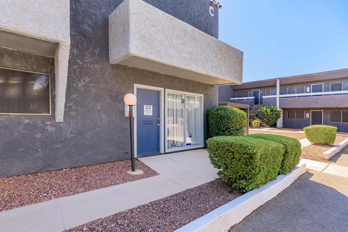 Exterior view of an apartment building featuring a gray facade and a blue front door with a window. A well-trimmed bush is located beside the entrance, and there is a light fixture on the left. The building is set in a landscaped courtyard with gravel and other units visible in the background.