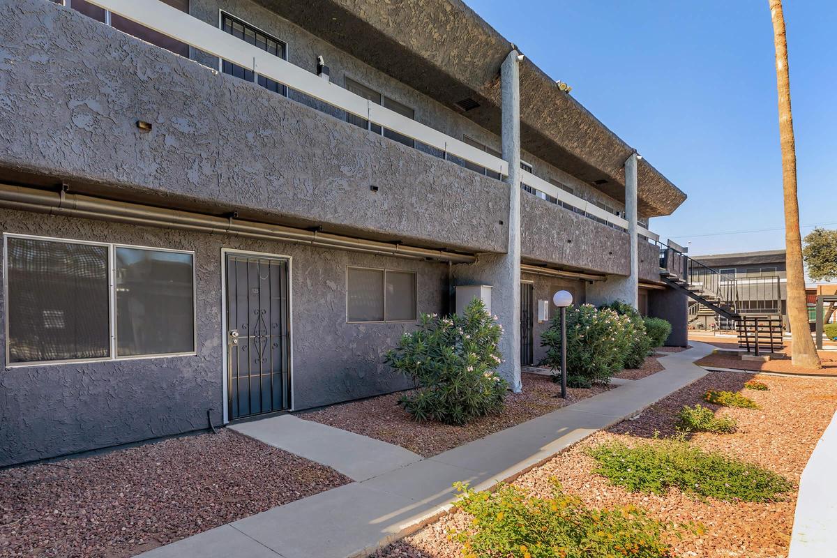 Exterior view of a multi-level apartment building with a textured gray façade. There are landscaped areas with shrubs and small plants along a paved walkway. A decorative lamp post and a staircase leading to the upper floor are visible. Clear blue sky in the background adds to the bright atmosphere.