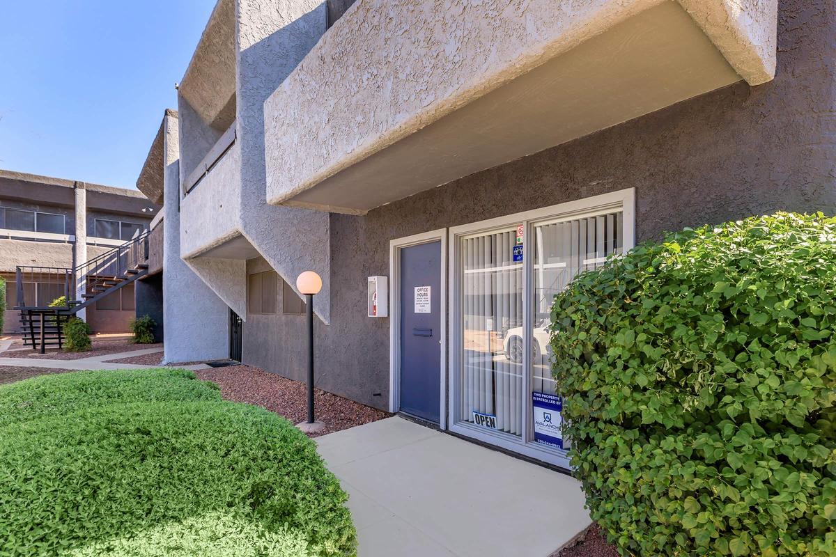 A modern apartment entrance featuring a gray wall with a door framed by a window. A potted bush is in front, and a lamp post stands nearby. The building has a textured exterior with a stairway visible in the background, reflecting a well-kept outdoor space.