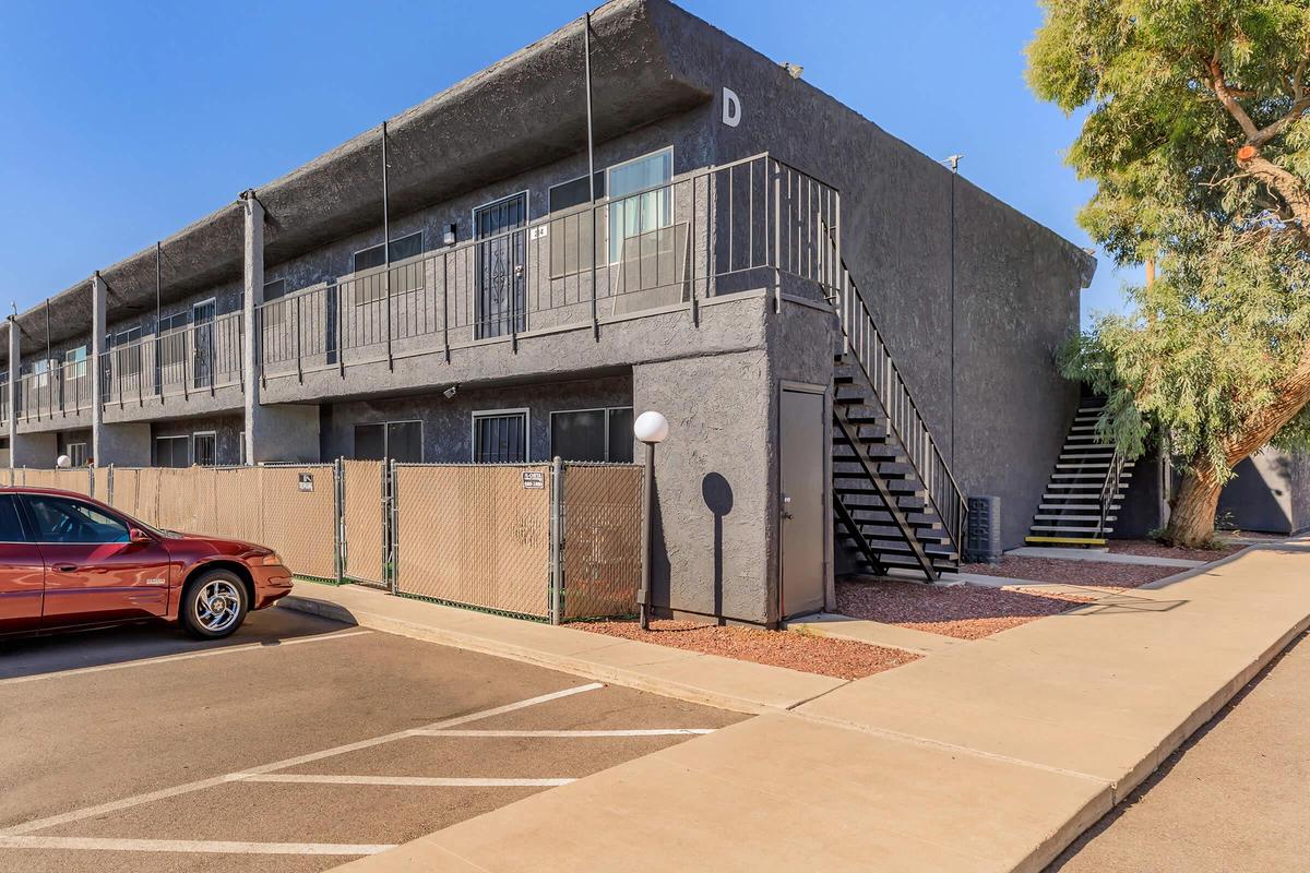 Two-story apartment building with a gray exterior. The building has multiple units with balconies and staircases on one side. In the foreground, there's a red car parked in front of a fenced area. A well-maintained walkway leads up to the building, surrounded by gravel and landscaping. Clear blue sky overhead.