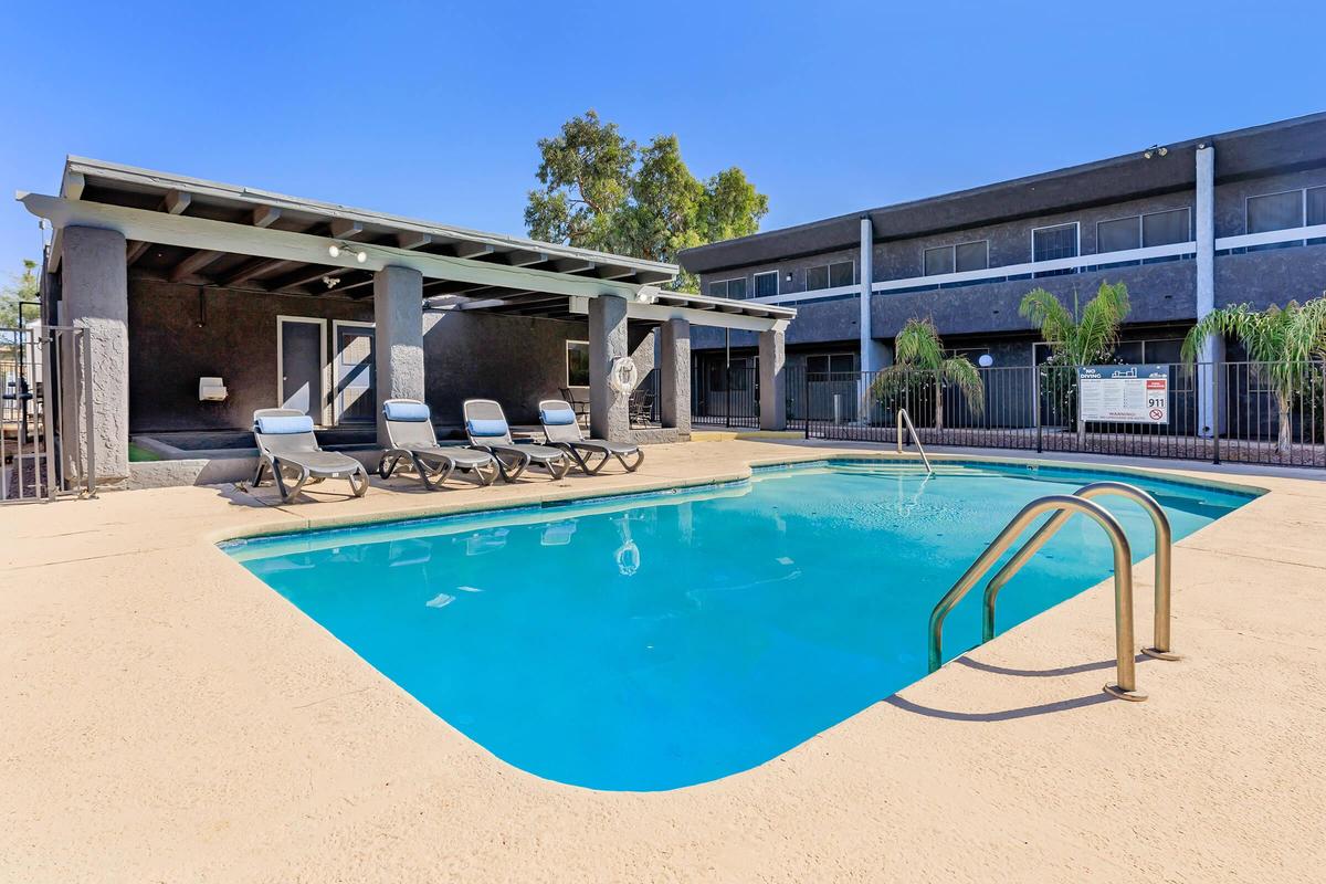 A sparkling swimming pool surrounded by lounge chairs, with a shaded seating area nearby. The pool area is well-maintained, featuring palm plants and a clean, inviting atmosphere. In the background, a multi-story building partially obscured by trees adds to the serene setting.