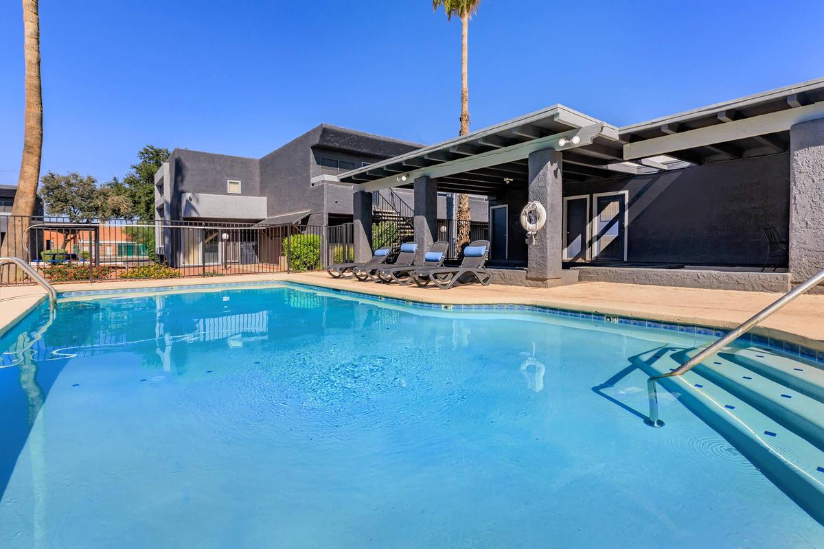 A sparkling blue swimming pool surrounded by lounge chairs, with a palm tree in the background. The pool area features a covered seating space and a fence. The sky is clear and sunny, creating a relaxing atmosphere.