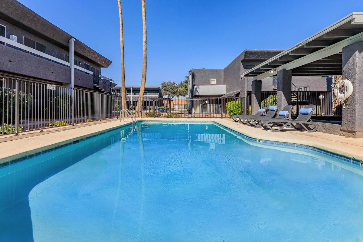 A clear blue swimming pool with lounge chairs beside it, surrounded by a fenced area. In the background, there are two-story buildings and palm trees under a bright blue sky. The scene conveys a relaxing outdoor space for leisure and enjoyment.